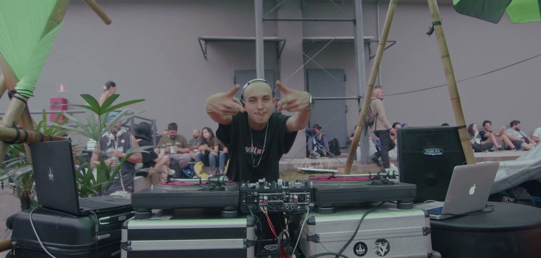 DJ tocando em set ao ar livre na área externa da feira, com público ao fundo