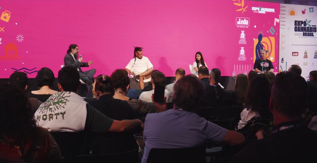 Debate no palco da ExpoCannabis Brasil, com o telão rosa e o público sentado em primeiro plano