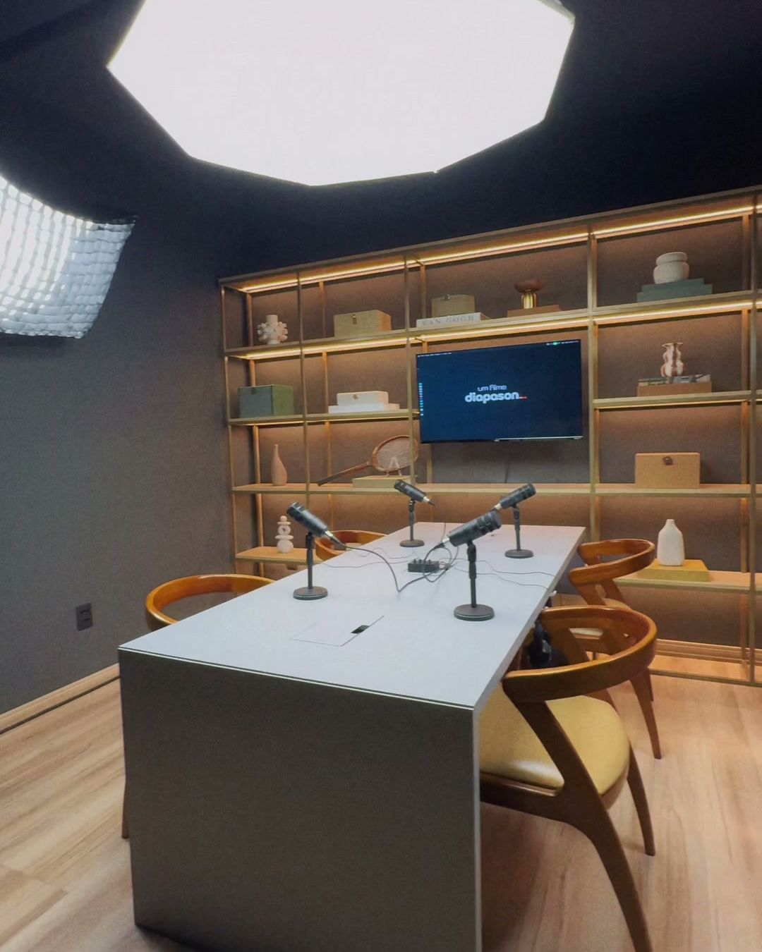 Estante iluminada e TV do Estúdio B · M8 vistas em grande angular, com a mesa de podcast em primeiro plano