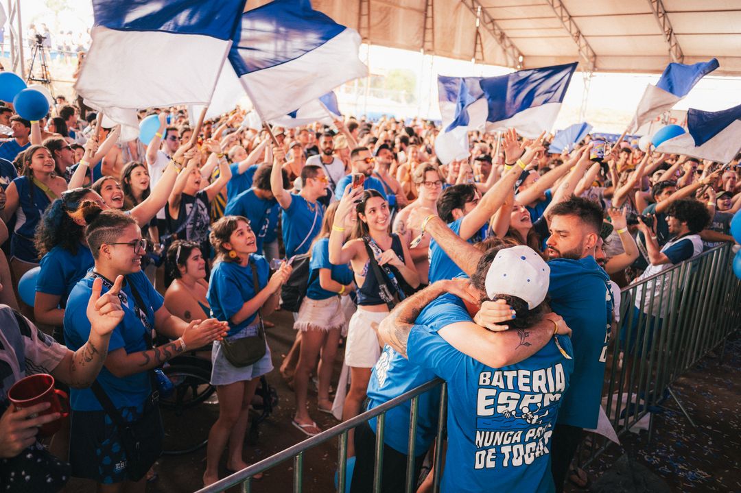 Torcida universitária de azul com bandeiras e braços erguidos durante a apresentação de uma bateria na Tabu 2025