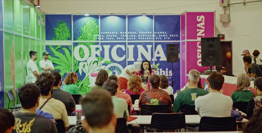 Oficina com plateia sentada em frente a um estande com o painel Oficinas