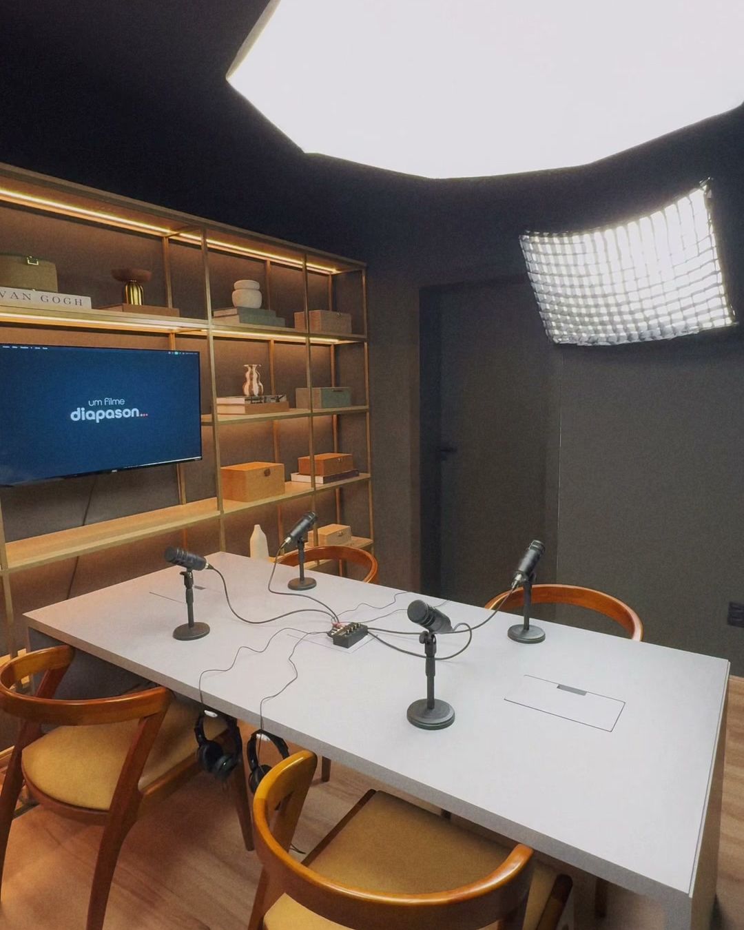 Set de podcast do Estúdio B · M8 visto de canto, com mesa branca, cadeiras, softbox octogonal e a porta acústica ao fundo